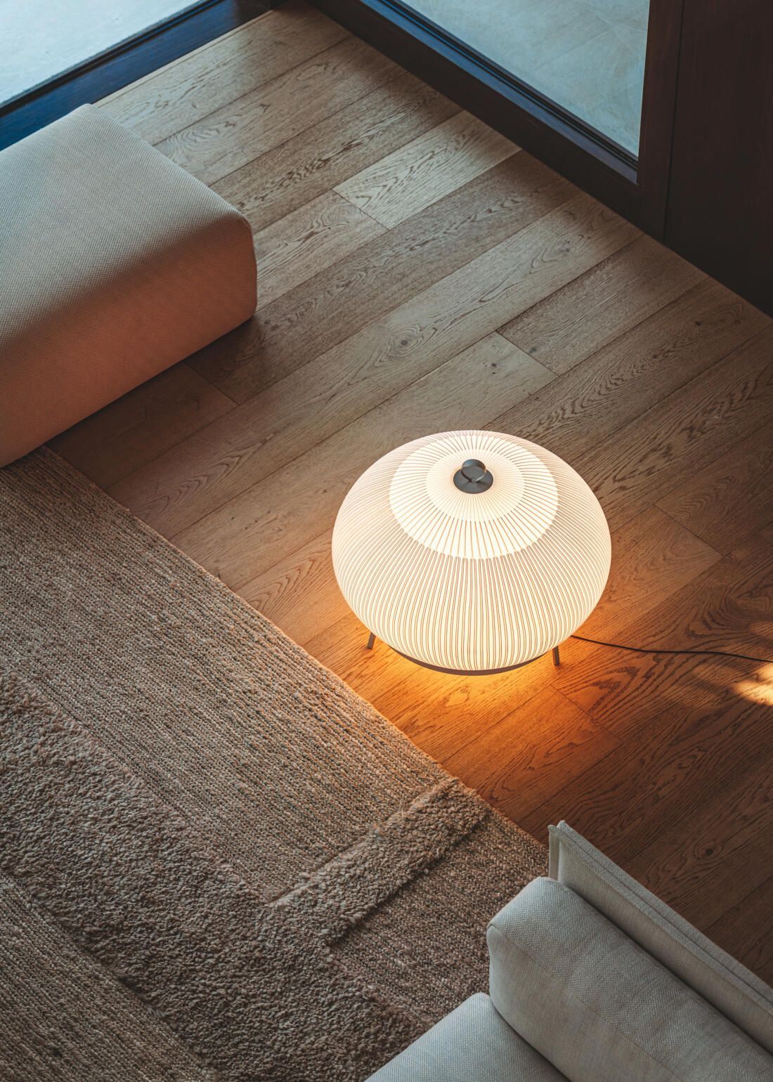 En una habitación minimalista, una lámpara esférica brillante con textura se encuentra sobre un suelo de madera, junto a una alfombra y un mueble.