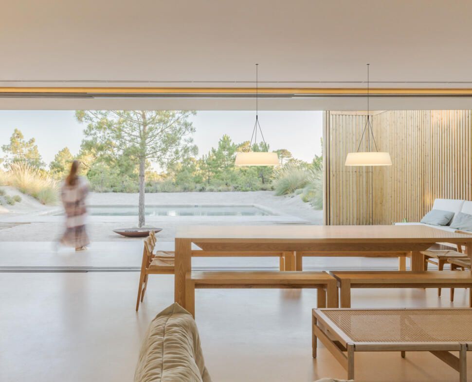 Comedor interior minimalista con muebles de madera, lámparas colgantes y una pared de cristal que se abre a una luminosa vista al jardín.