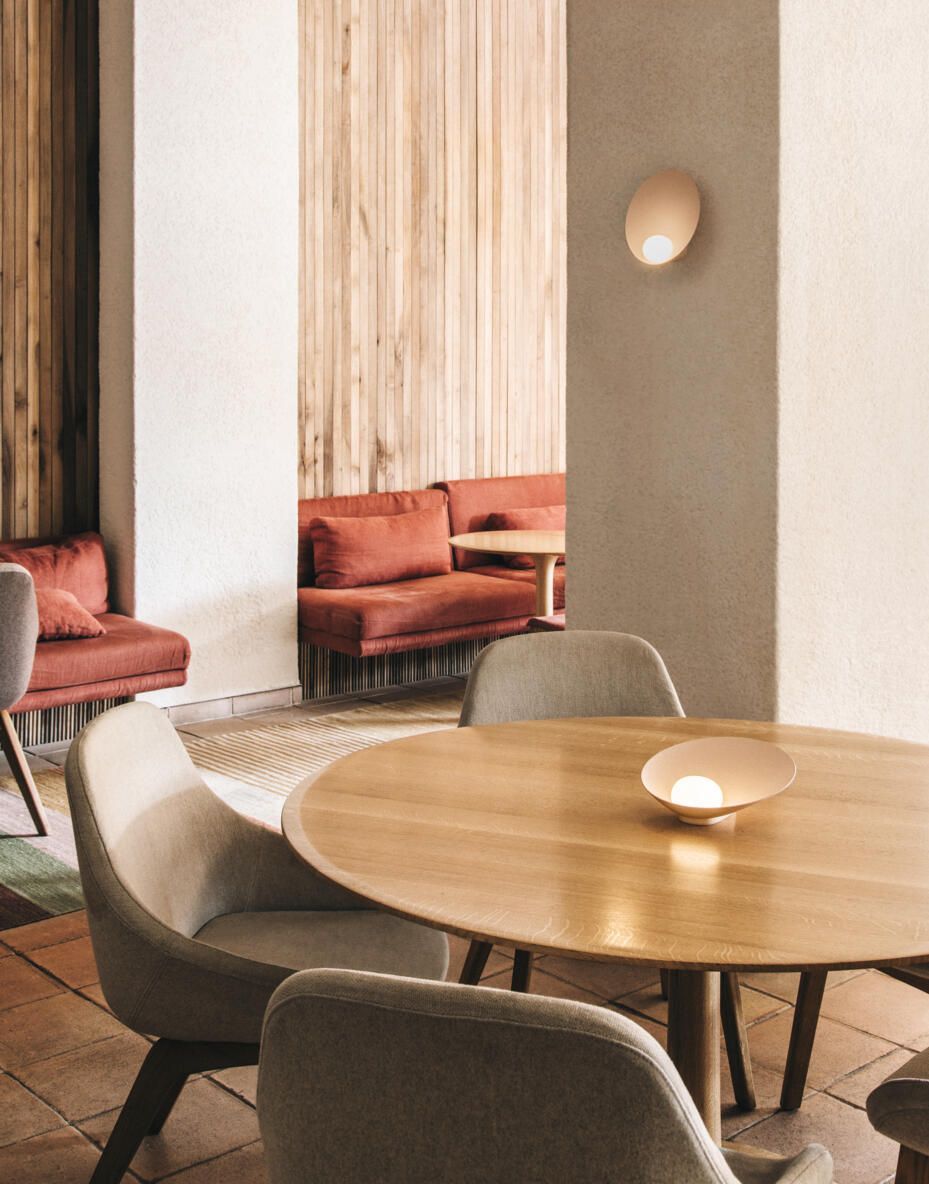 El interior de una cafetería moderna cuenta con una mesa redonda de madera, sillas tapizadas en gris y bancos de color terracota.
