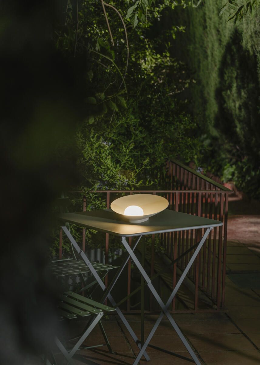 Por la noche, una lámpara de mesa encendida, con forma de cuenco, reposa sobre una pequeña mesa de exterior, rodeada de exuberante vegetación.