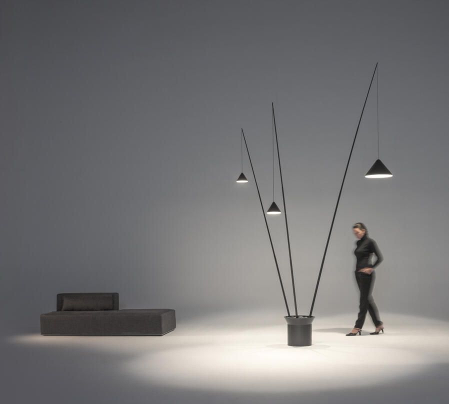 Una lámpara de pie moderna con tres brazos altos y delgados que sostienen luces colgantes, junto a un sofá bajo y una persona que pasa caminando.