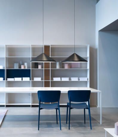Dos sillas azules se encuentran frente a una mesa blanca, delante de una gran estantería blanca con dos lámparas colgantes cónicas negras.