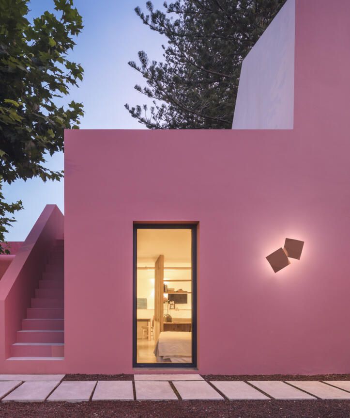 Una pared exterior de color rosa cuenta con una puerta de cristal vertical, una escalera exterior y dos apliques de pared cuadrados que emiten luz.