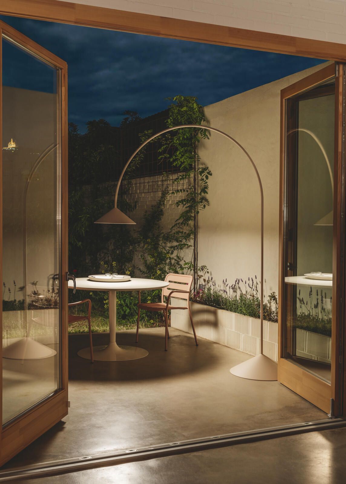 Un comedor al aire libre al atardecer, con una mesa circular, sillas y una lámpara de pie alta y arqueada, enmarcado por puertas de madera.