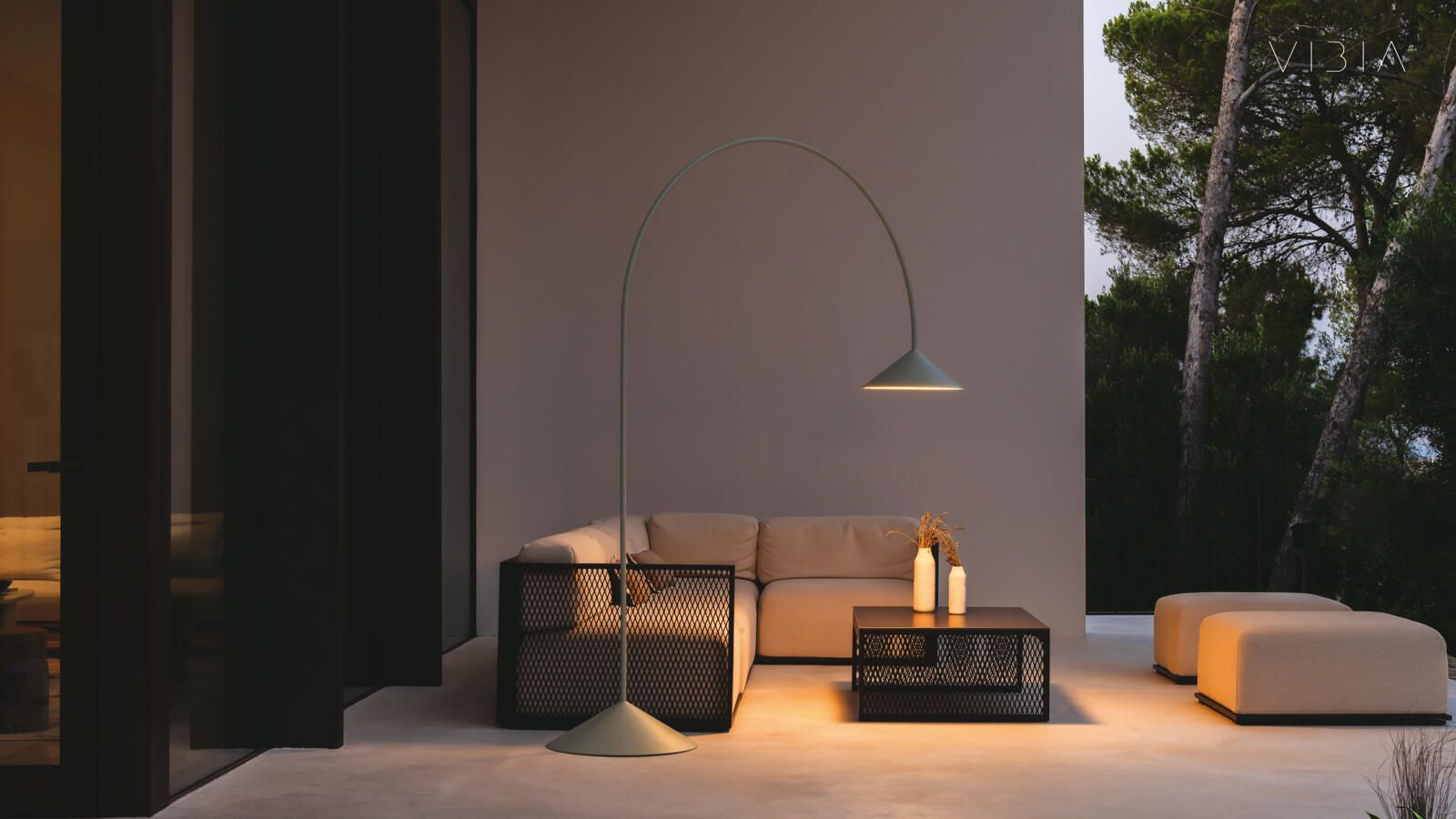 Una zona de estar exterior minimalista con una elegante lámpara de pie curvada, un sofá beige, una mesa auxiliar estampada y dos otomanas.