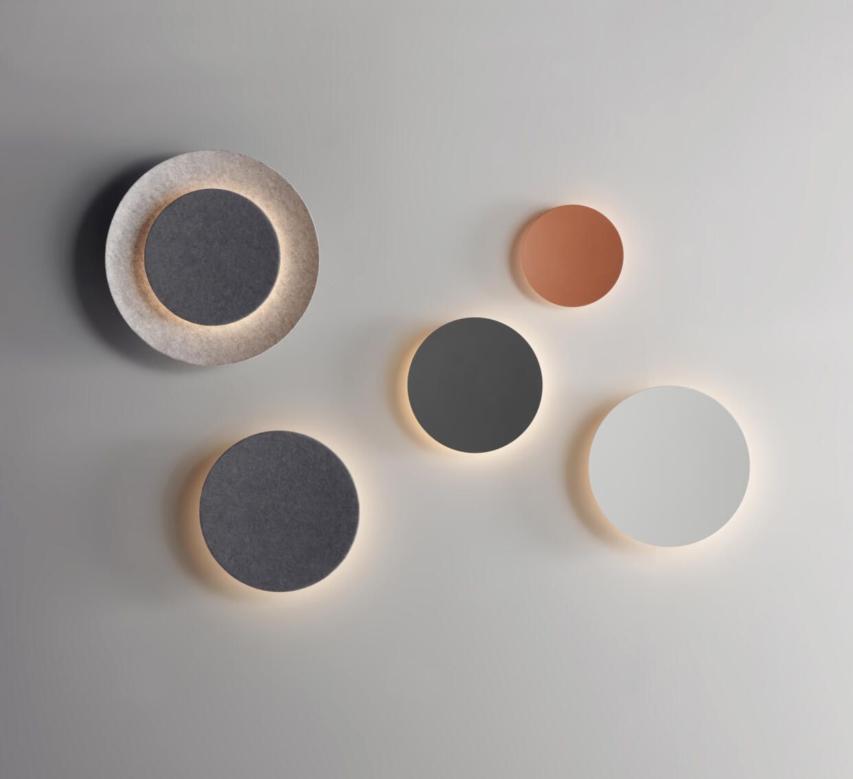 Cinco apliques circulares de pared de diferentes tamaños y colores (gris, blanco y terracota) dispuestos sobre una pared lisa.