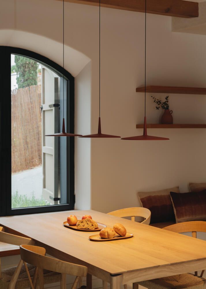 Una mesa de comedor de madera con pan en una habitación luminosa y moderna con ventanas arqueadas, iluminación minimalista y estantes en la pared.