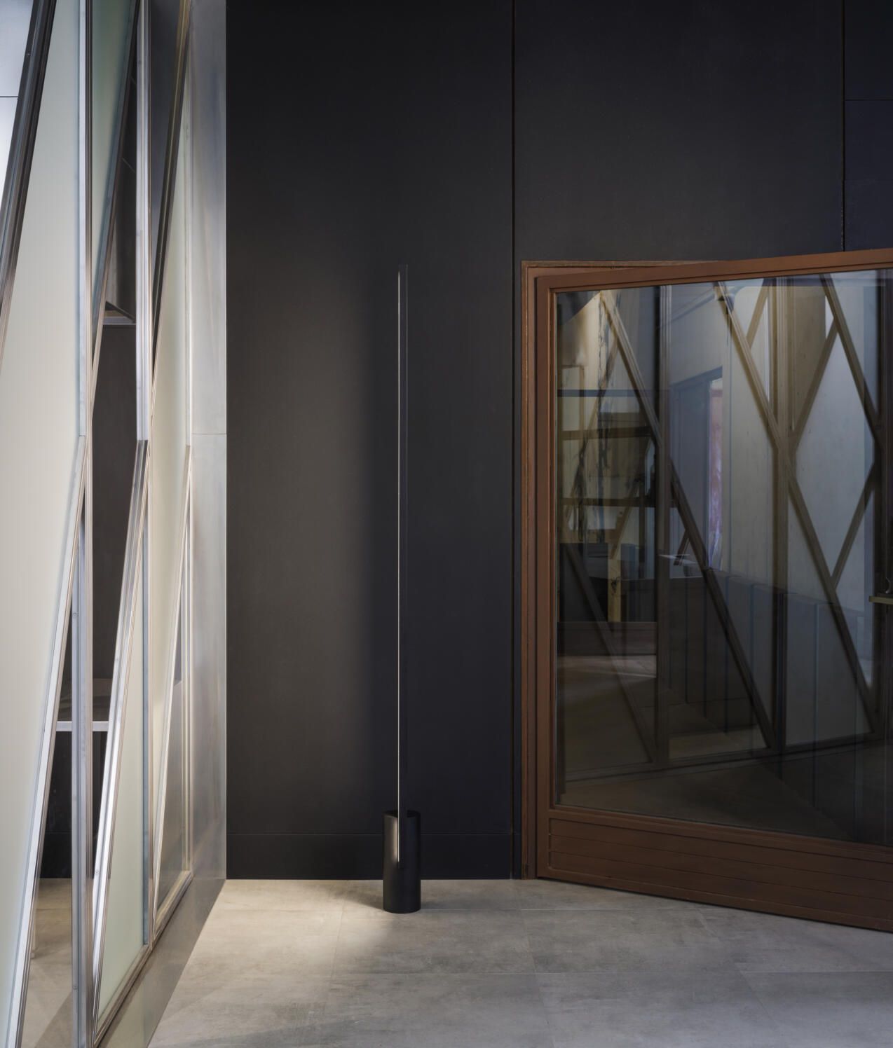 Una lámpara de pie alta y esbelta se alza en un rincón, contra una pared oscura, junto a una puerta con un diseño geométrico de cristal.