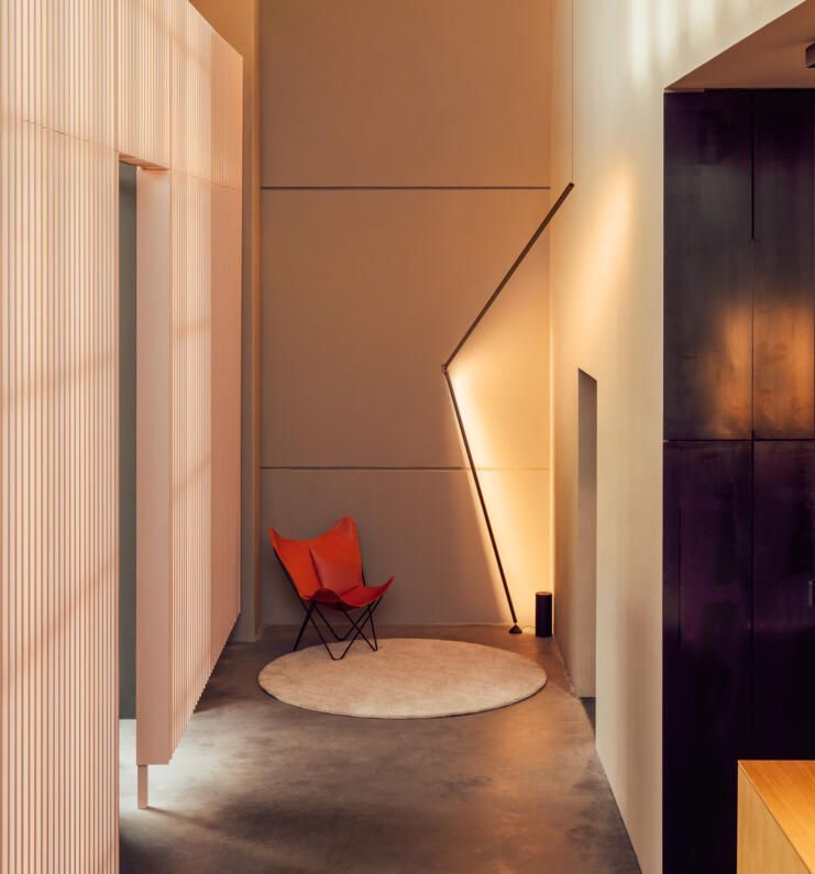 Una silla mariposa de color naranja reposa sobre una alfombra redonda en un pasillo moderno iluminado por una lámpara de pie angular y de diseño llamativo.