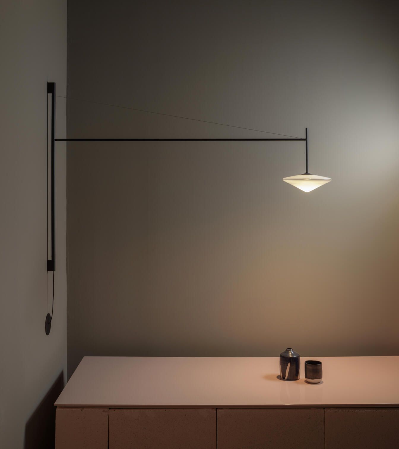 Una lámpara de pared minimalista con un brazo negro delgado y una pantalla brillante en forma de disco, colocada sobre una mesa con objetos.