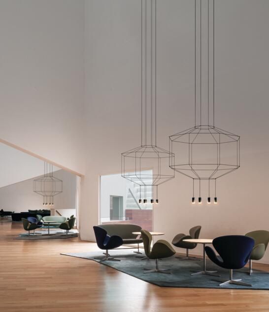 Un salón luminoso y moderno con lámparas colgantes de cable visto sobre conjuntos de sillones y mesas dispuestas sobre una gran alfombra.