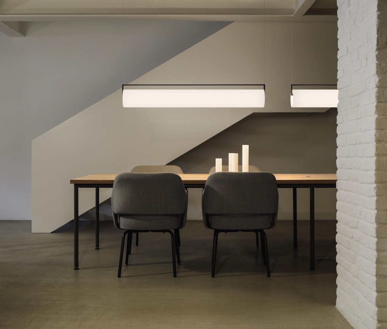 Un comedor moderno con dos sillas grises, una mesa larga de madera, velas cilíndricas encendidas y dos lámparas colgantes lineales.