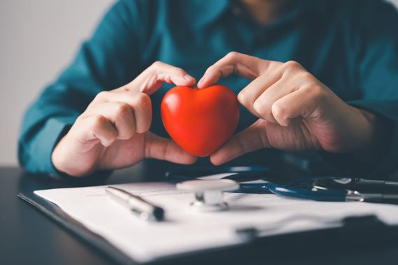 Hands held in a heart shape framing a small red heart model, resting on a document with a stethoscope and pen.
