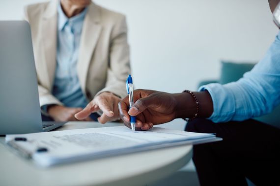 A person in a blue shirt signs a document on a clipboard while another person points at it next to a laptop.