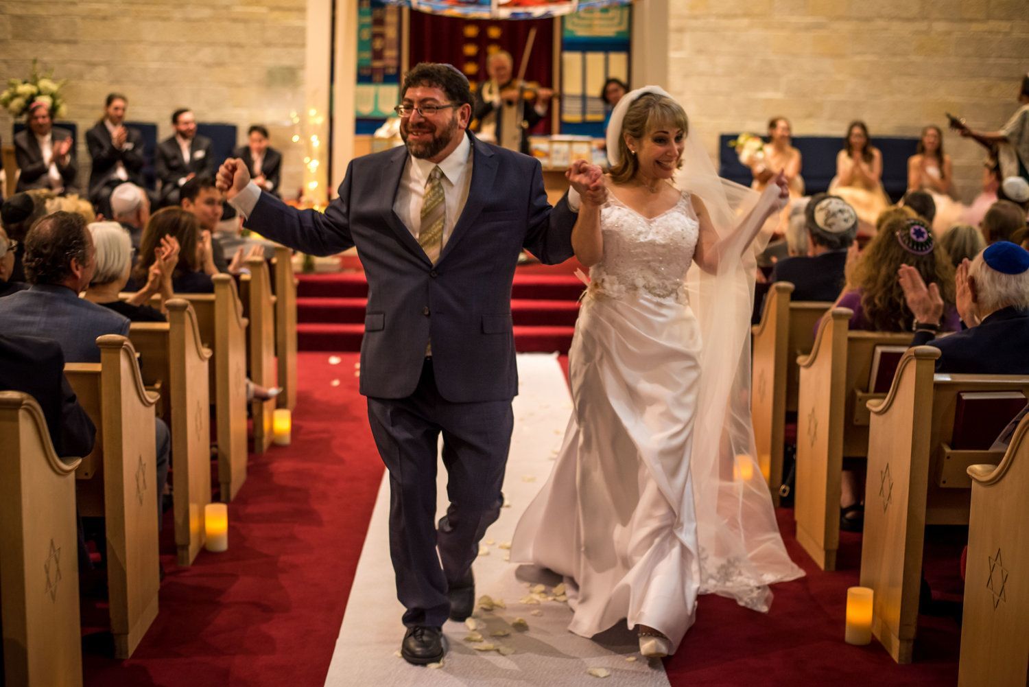 Newly married couple walking down the aisle, holding hands, smiling, surrounded by guests in a synagogue.