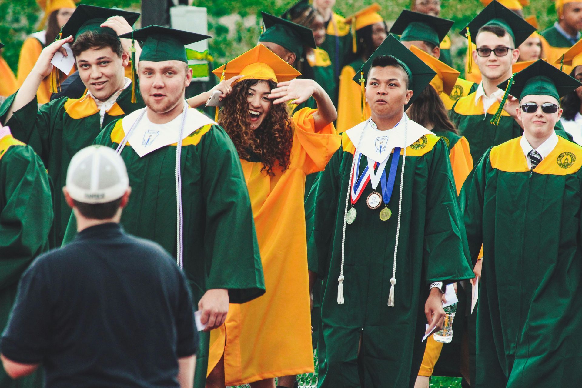 Graduates in green and gold gowns walk, some smiling, waving, or adjusting caps, at an outdoor ceremony.