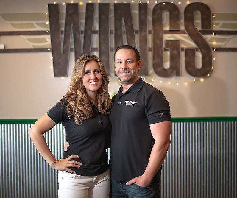 A man and a woman are posing for a picture in front of a sign that says wings