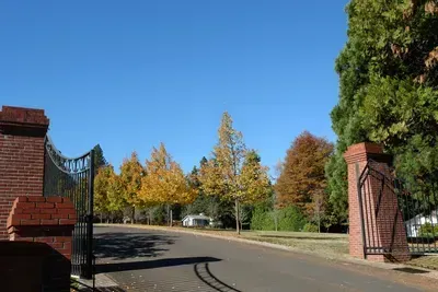 Open brick gate leading to a road lined with trees with yellow and orange leaves on a clear, sunny day.