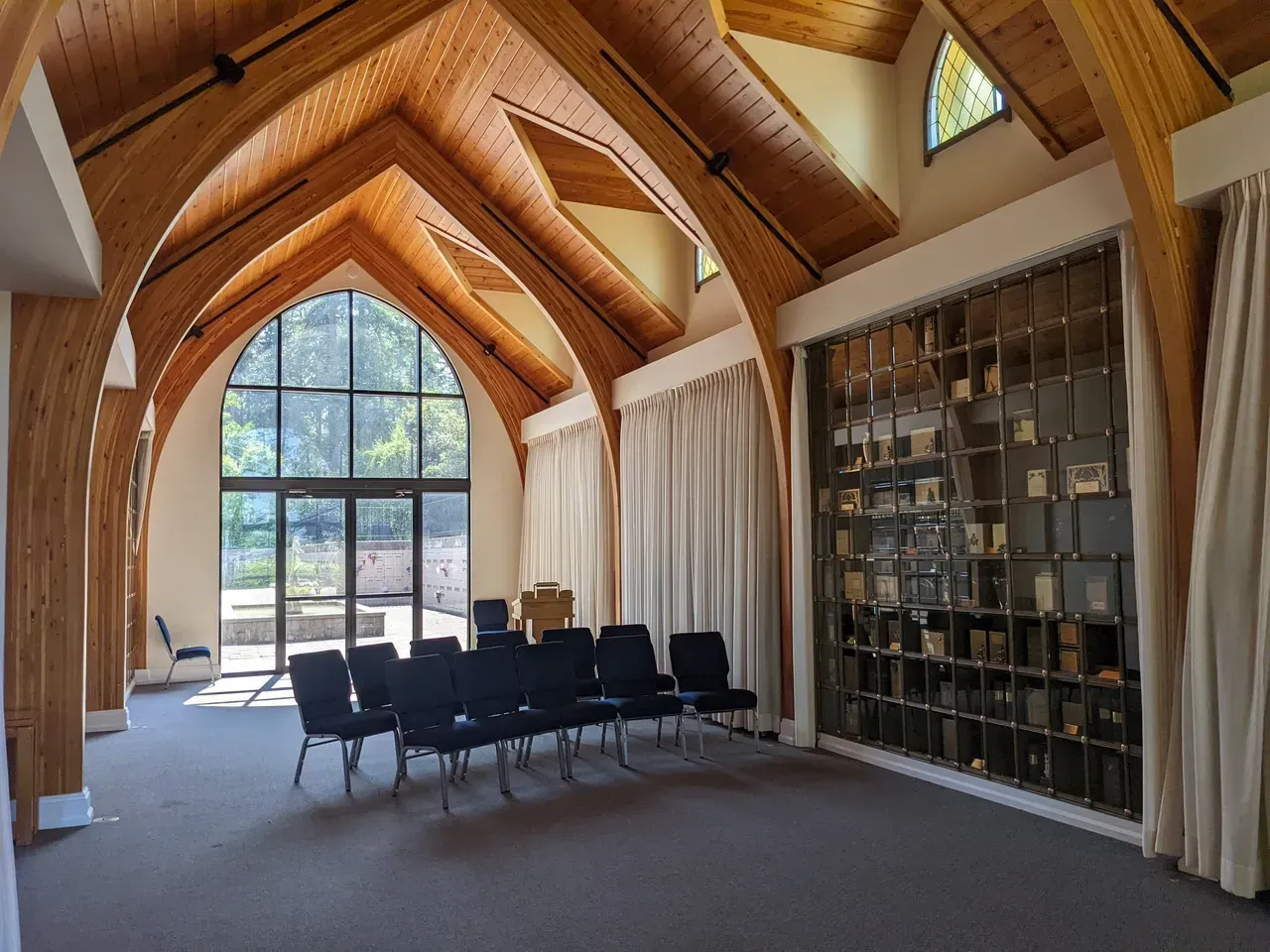 Interior of a chapel with high, wooden arched ceiling and glass-fronted columbarium wall; rows of chairs face large window.