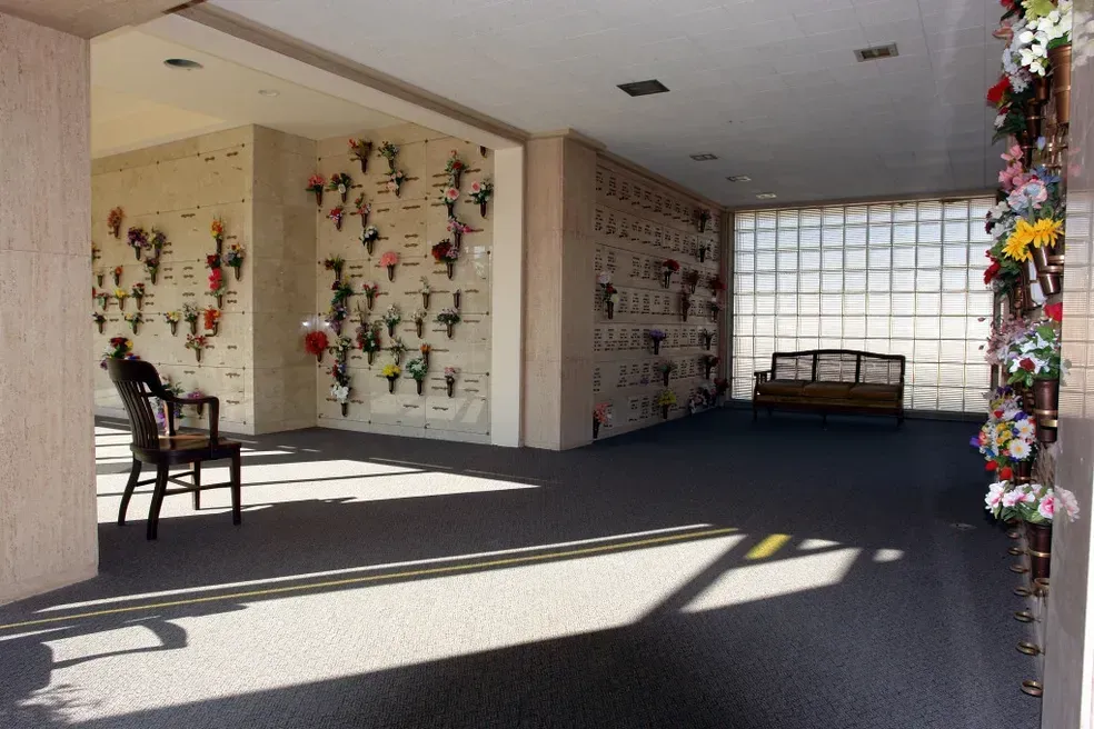 Interior of a mausoleum with niches holding urns and flowers. A chair and a bench sit in the space. Sunlight streams in.