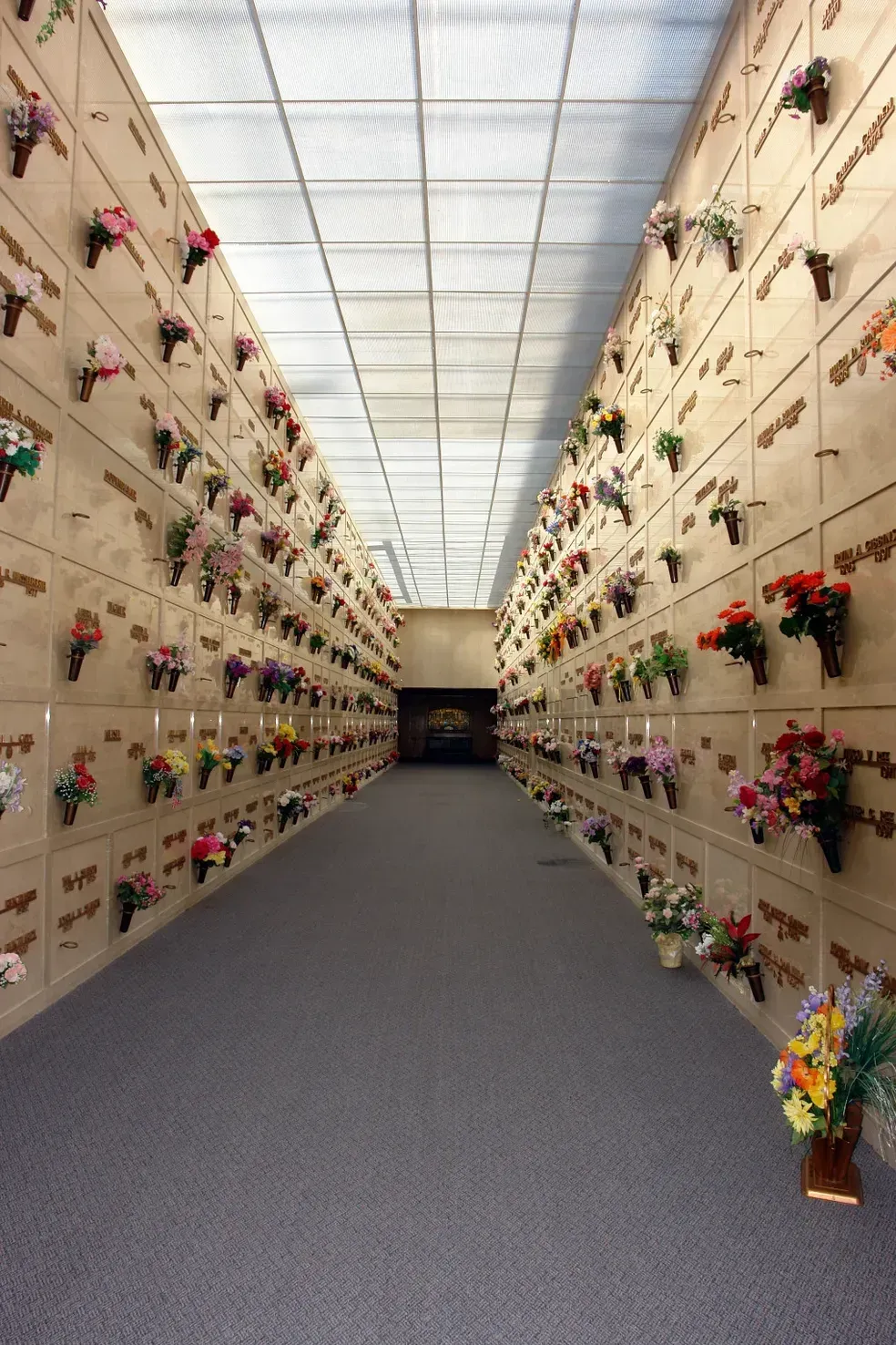Interior view of a mausoleum. Walls lined with niches, each with a vase of flowers, along a central gray carpeted aisle.