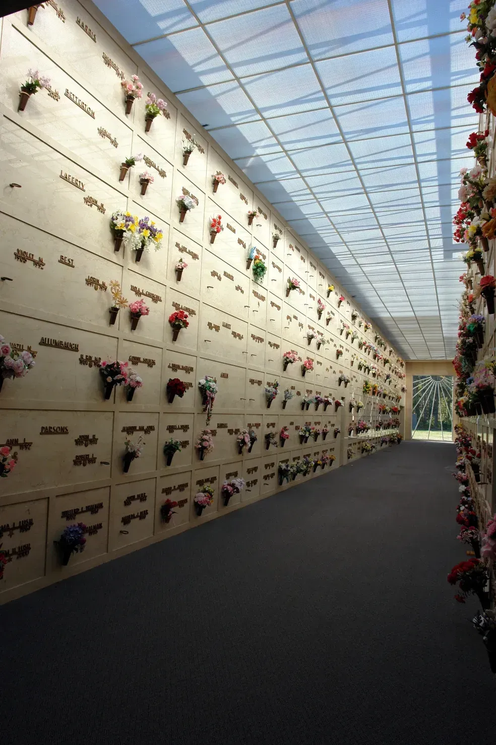 Interior of a columbarium with rows of niches holding urns, decorated with flowers. Sunlight streams through the skylight.