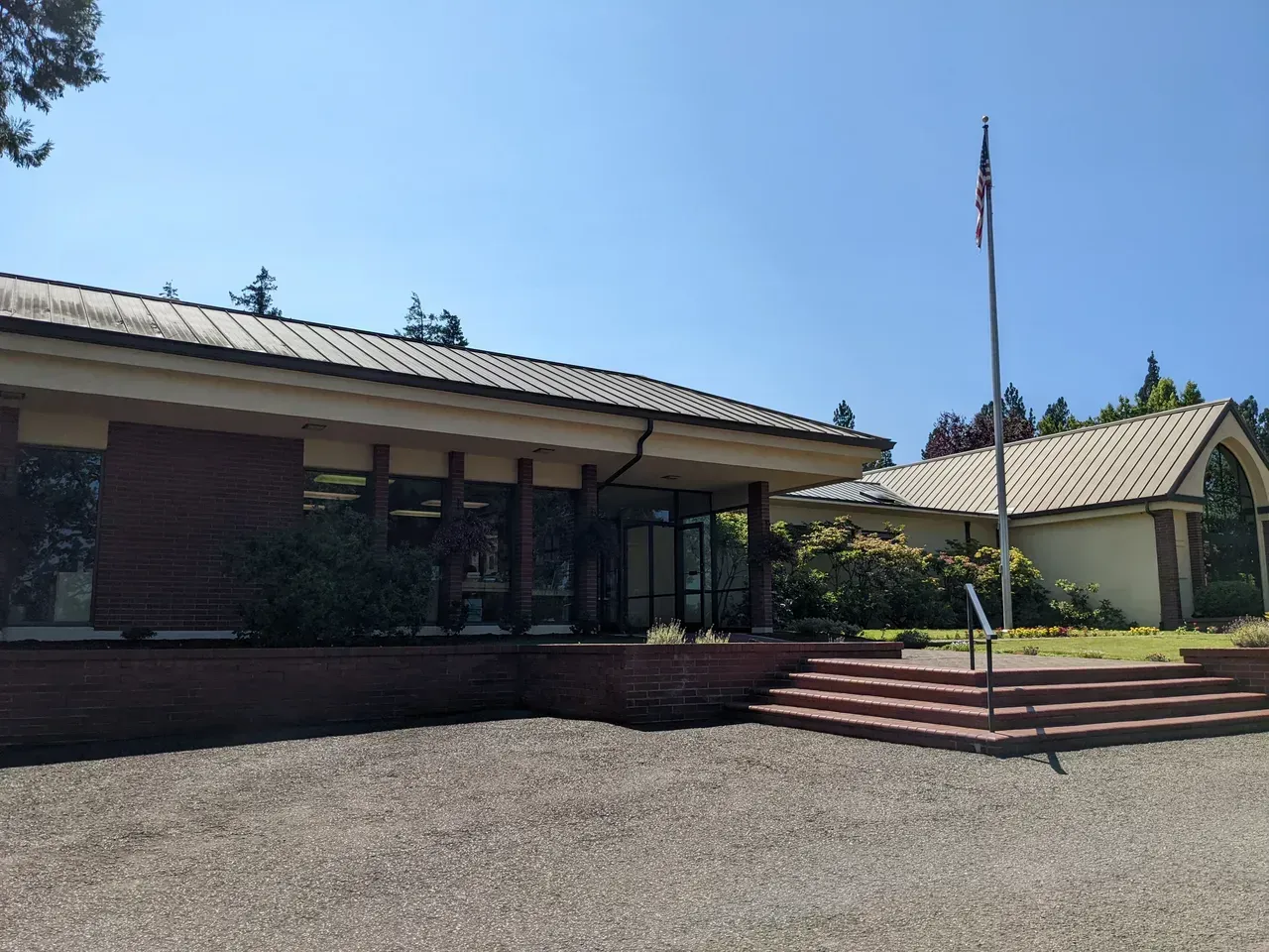 A low, single-story building with a brick facade and dark roof on a sunny day. A flag pole stands to the right.