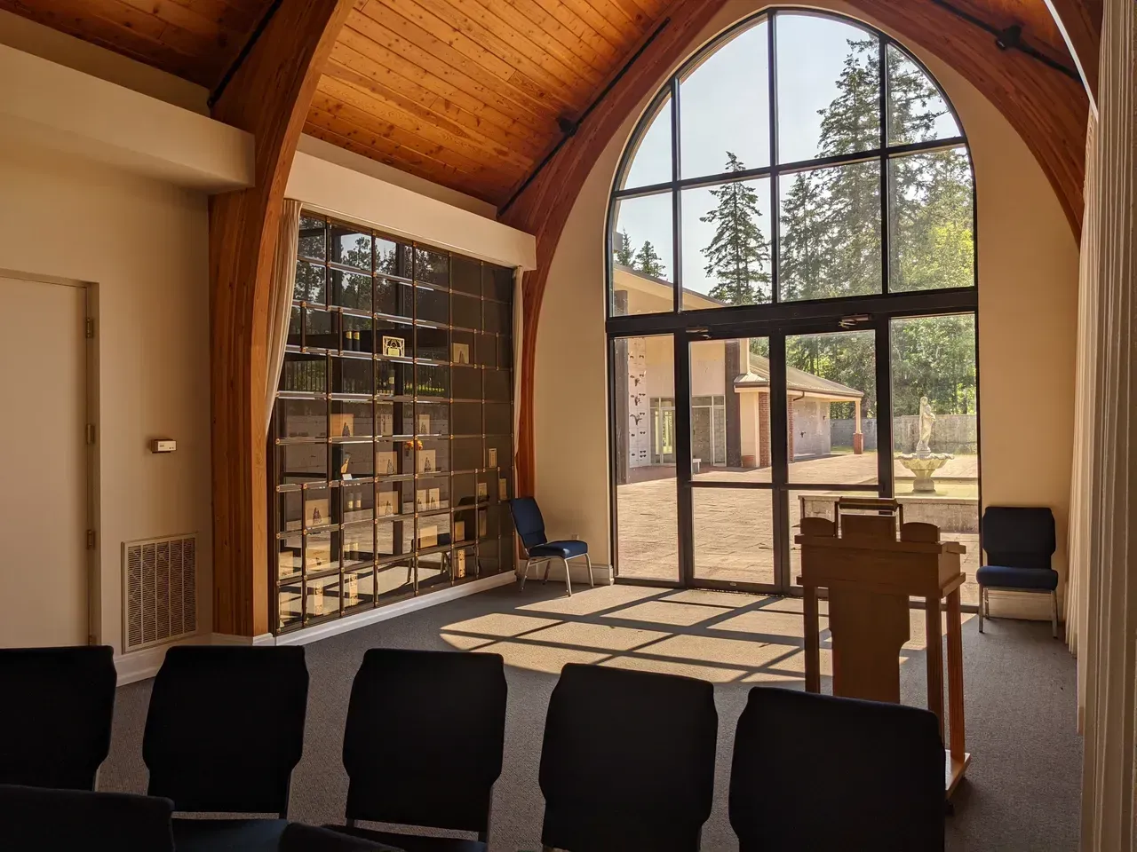 Interior chapel with large arched window, wooden ceiling, and rows of seating.