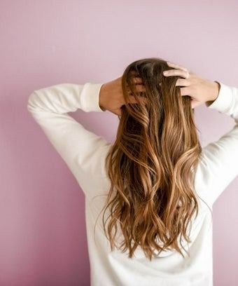 Person with long, wavy brown hair holding their head, wearing a white sweater, in front of a pink wall.