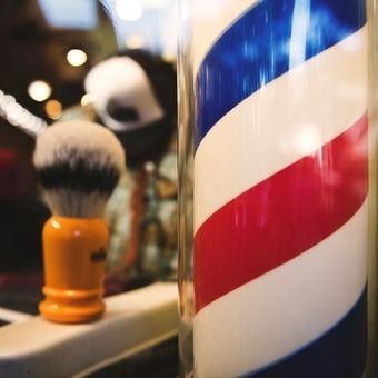 Barber pole with red, white, and blue stripes; shaving brush with orange handle; blurred background.