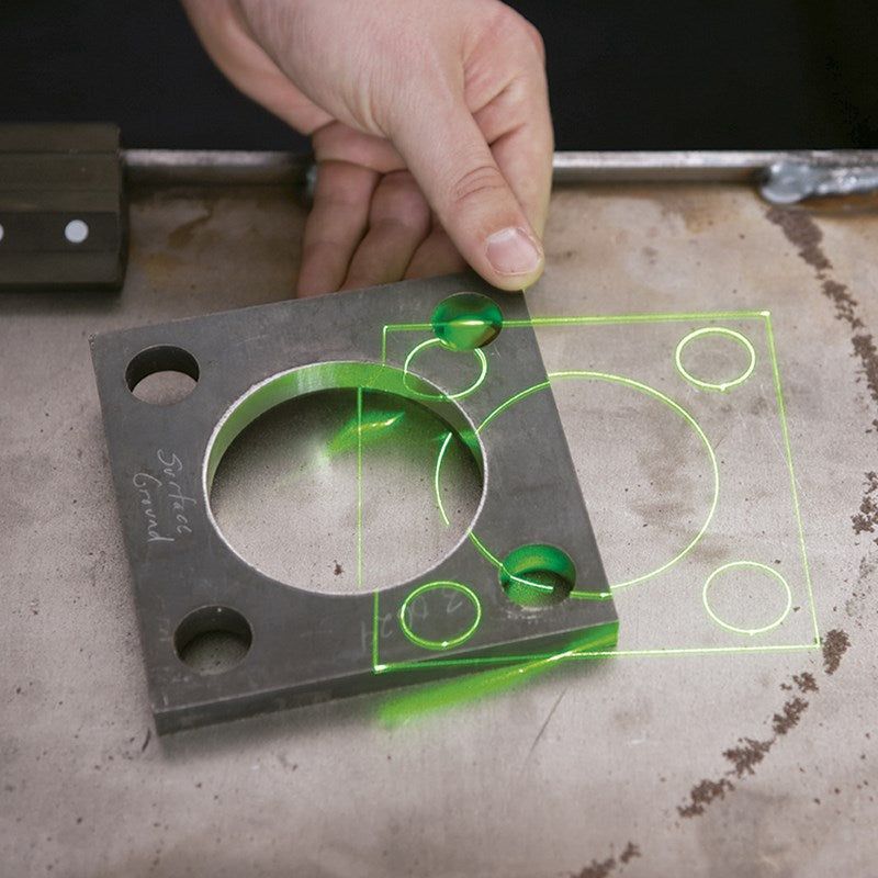 Hand holding a clear template over a metal plate, showing green laser alignment on a workbench.