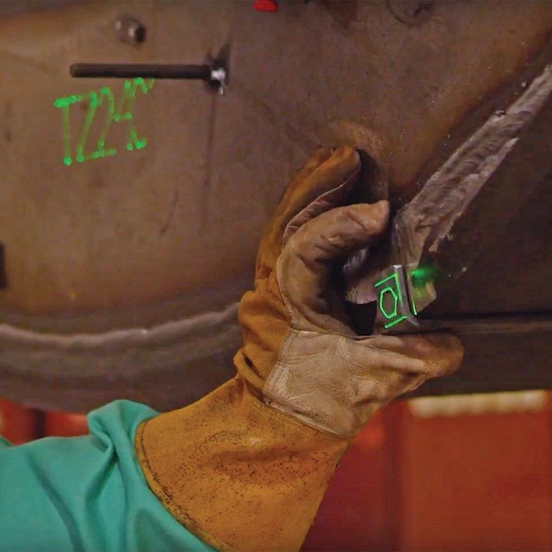 A gloved hand holds a device with a green laser, near a weld on metal.