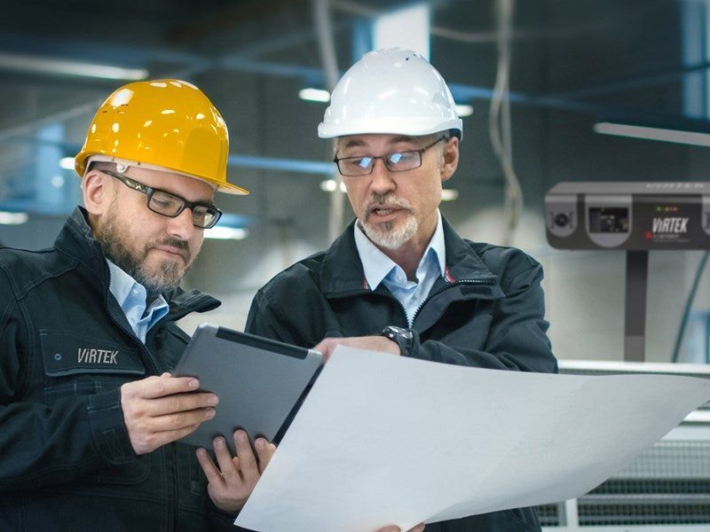 Two men in hard hats reviewing blueprints and a tablet in a factory setting.