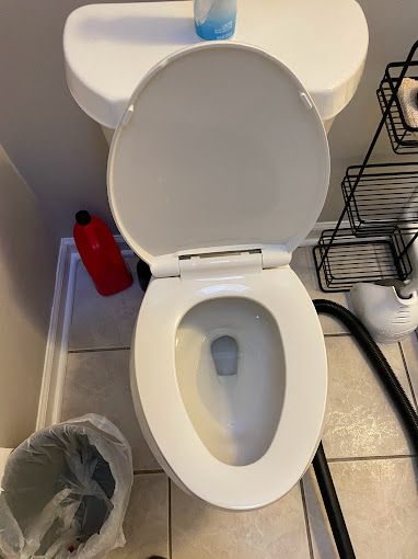 A white toilet with the lid up in a bathroom next to a vacuum cleaner.