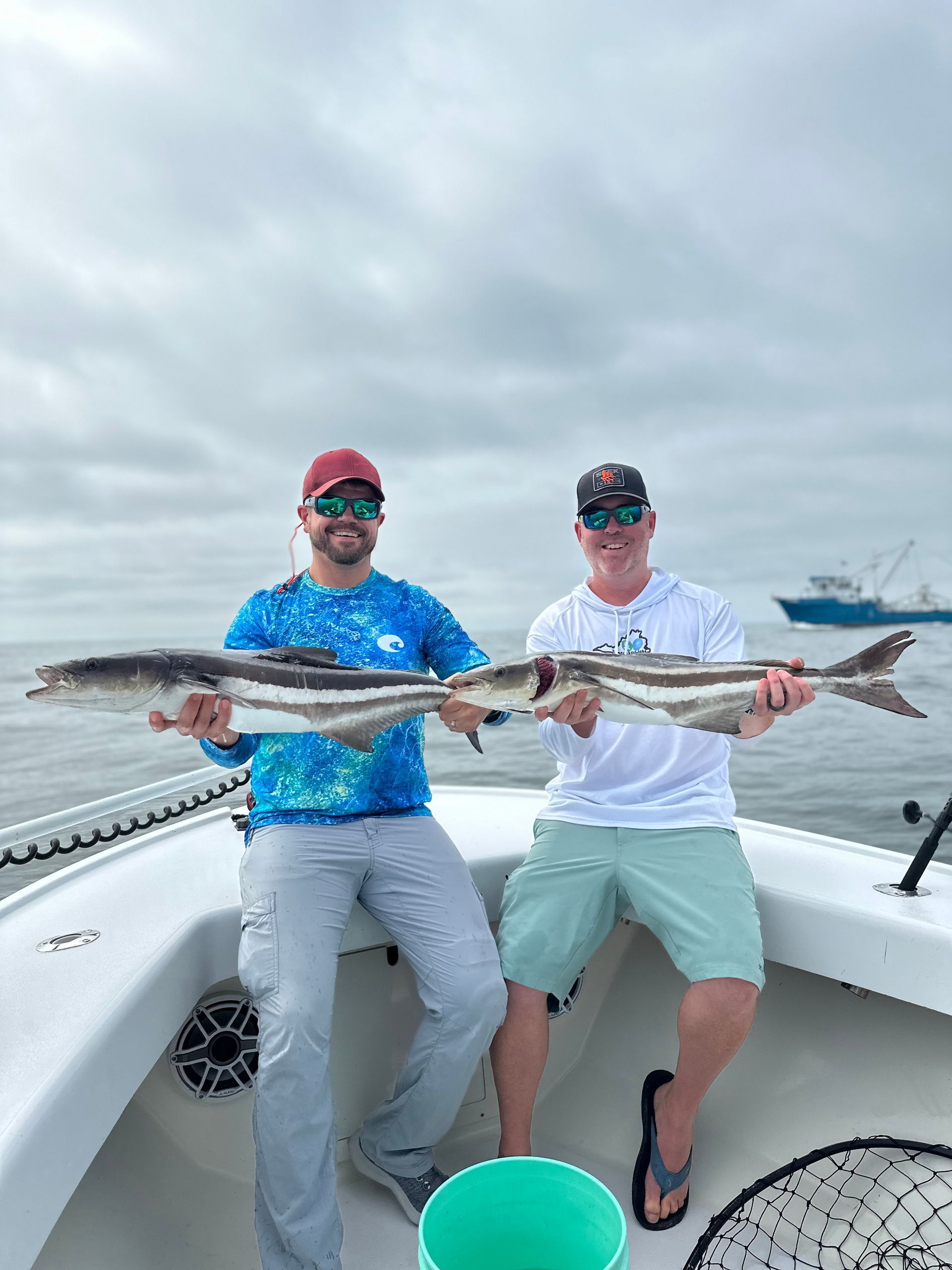 Two men are sitting on a boat holding two large fish.