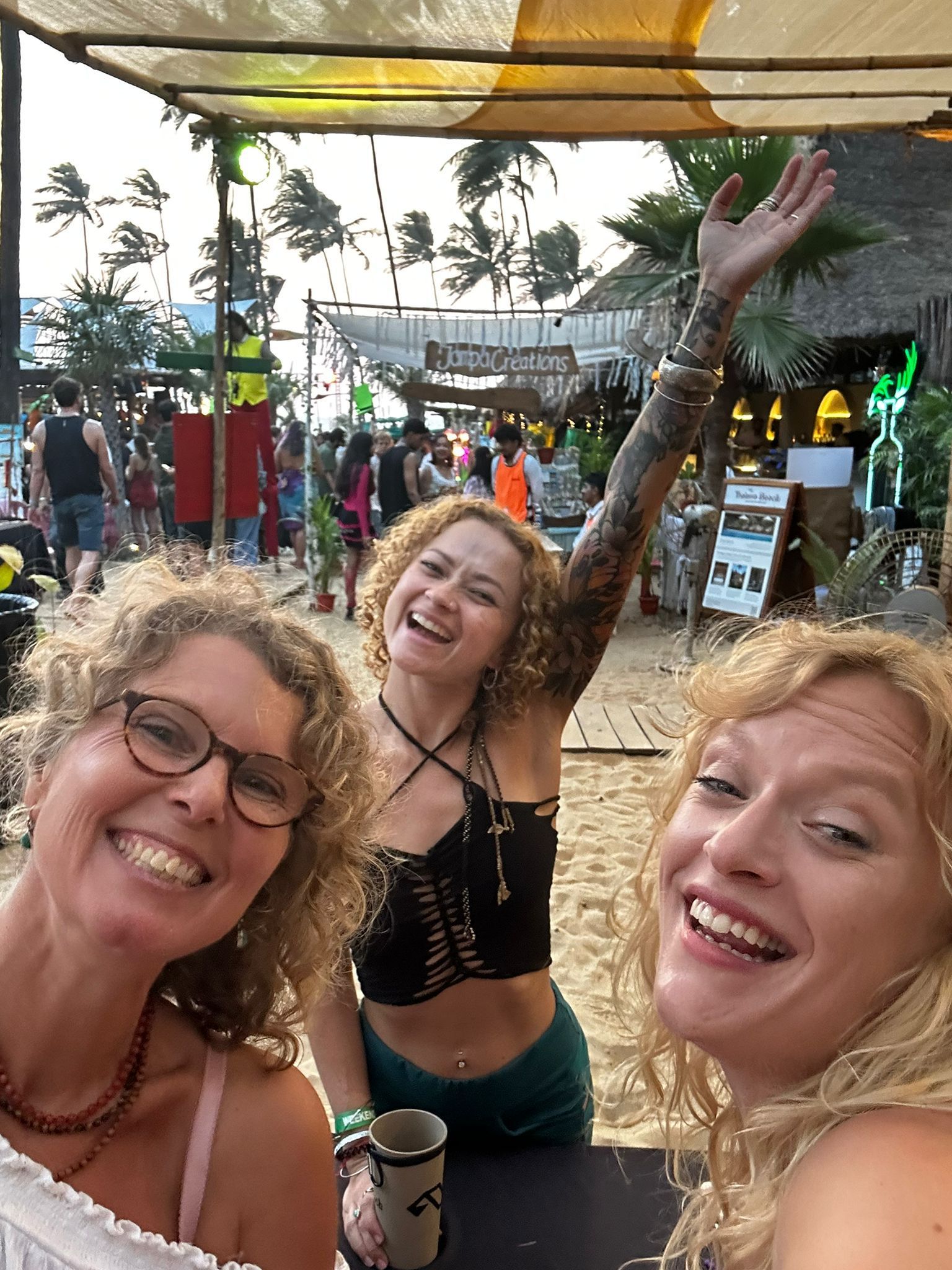 Three smiling women pose for a selfie at a beachside cafe; one waves with a full arm tattoo.