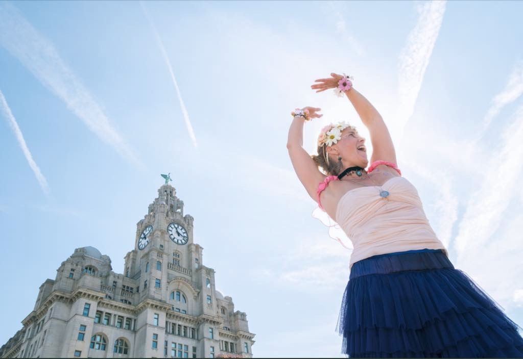 Woman in costume poses with arms raised, Liverpool's Royal Liver Building in background, blue sky.