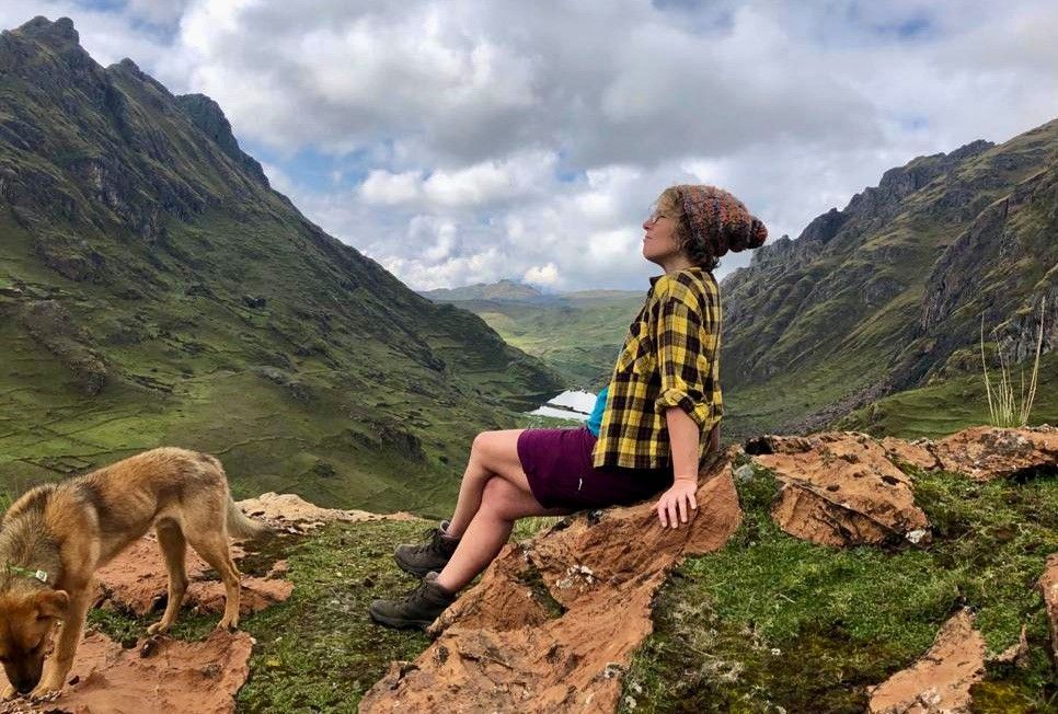 Woman sitting on a rock in a valley, looking up at the sky. A dog is nearby, mountains in background.