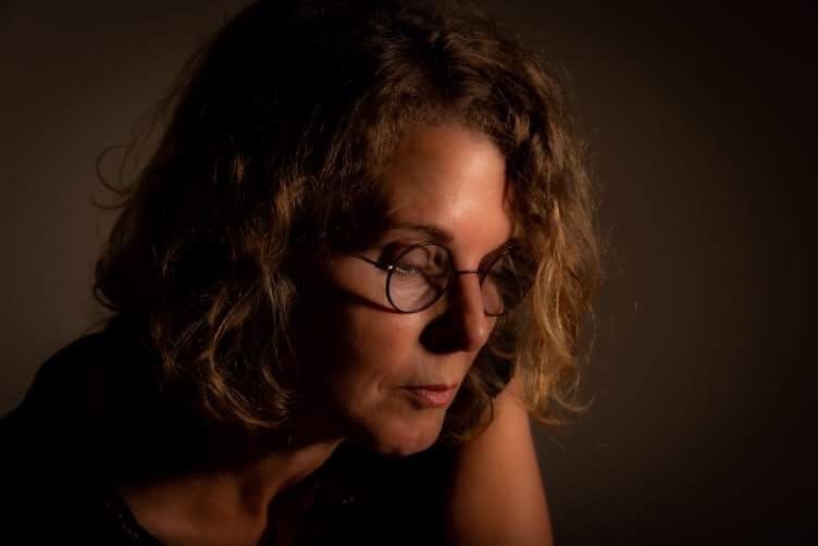 Woman wearing glasses, looking down thoughtfully with hand near face. Dark setting with focused lighting.