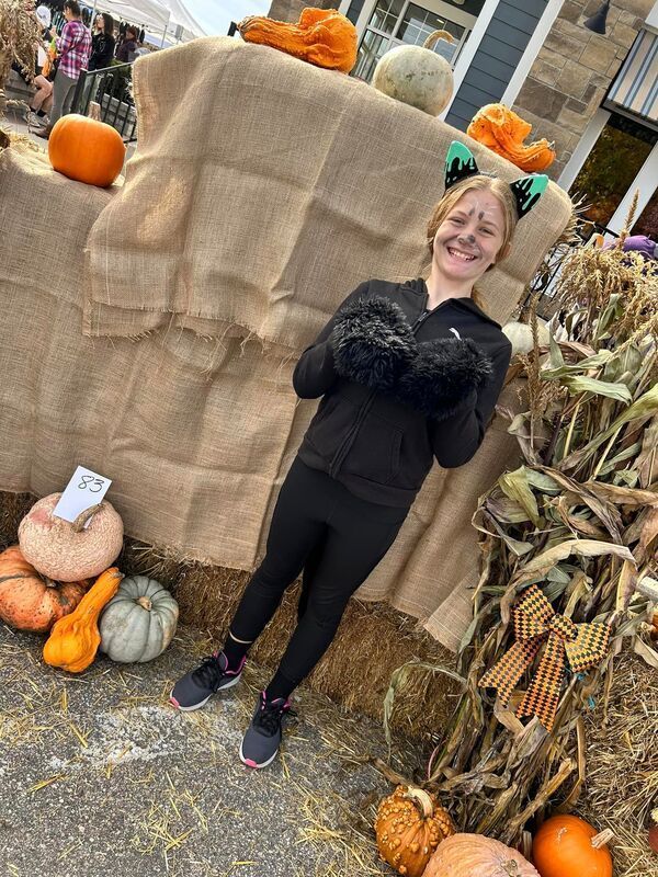 A woman in a cat costume is standing in front of a pile of pumpkins.