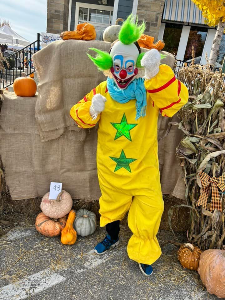 A clown in a yellow costume is standing in front of pumpkins.