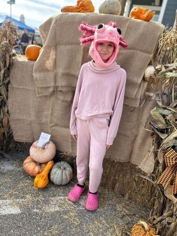A child wearing an axolotl hat is standing next to some pumpkins