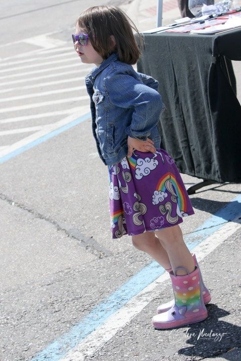 A little girl wearing a purple skirt and pink rain boots is standing in a parking lot.