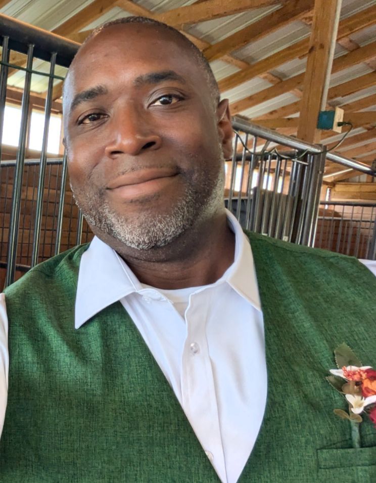 A smiling person wearing a green vest over a white collared shirt with a floral boutonnière, inside a wooden barn.