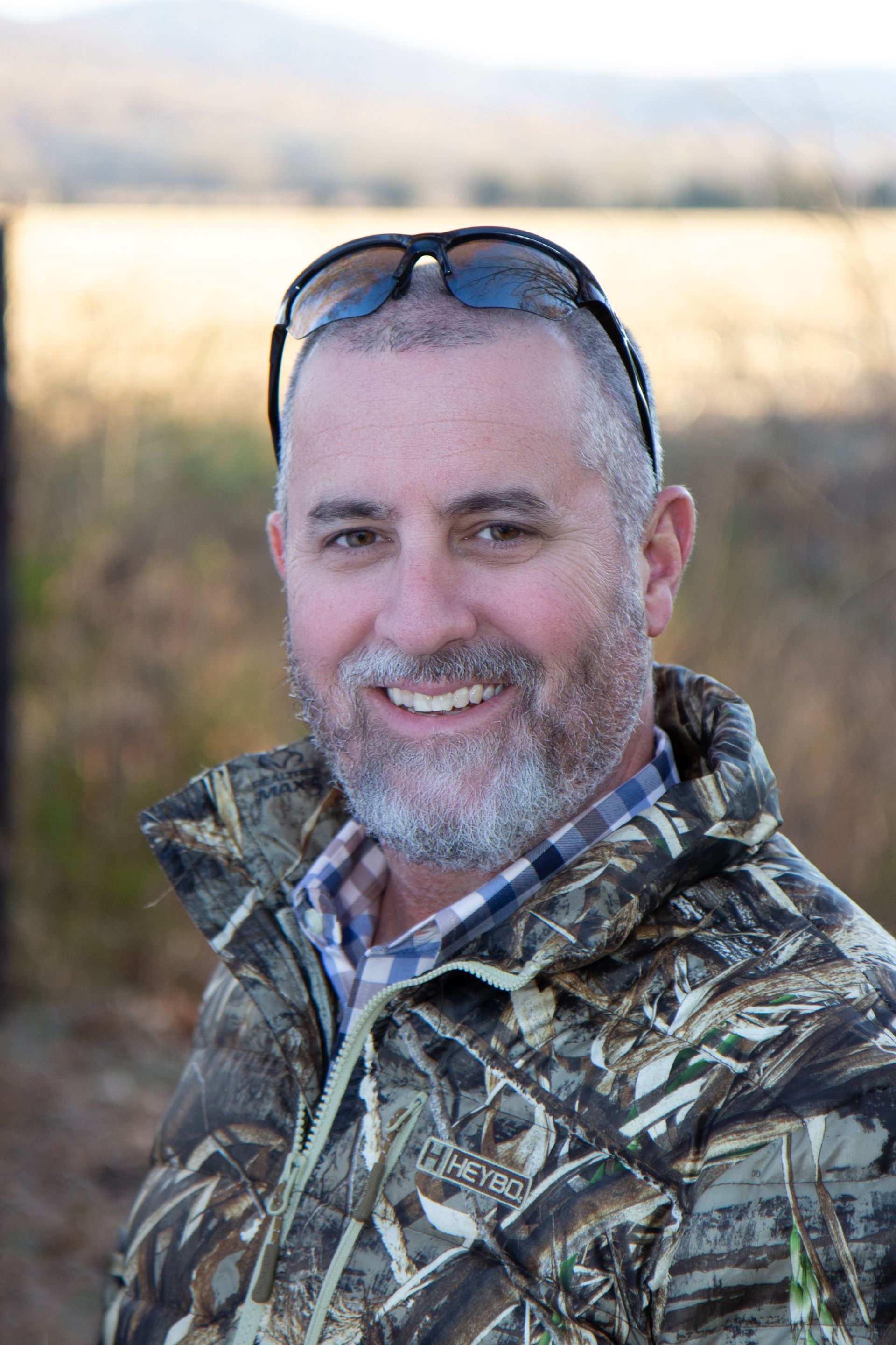 A man wearing a camouflage jacket and sunglasses is smiling for the camera.