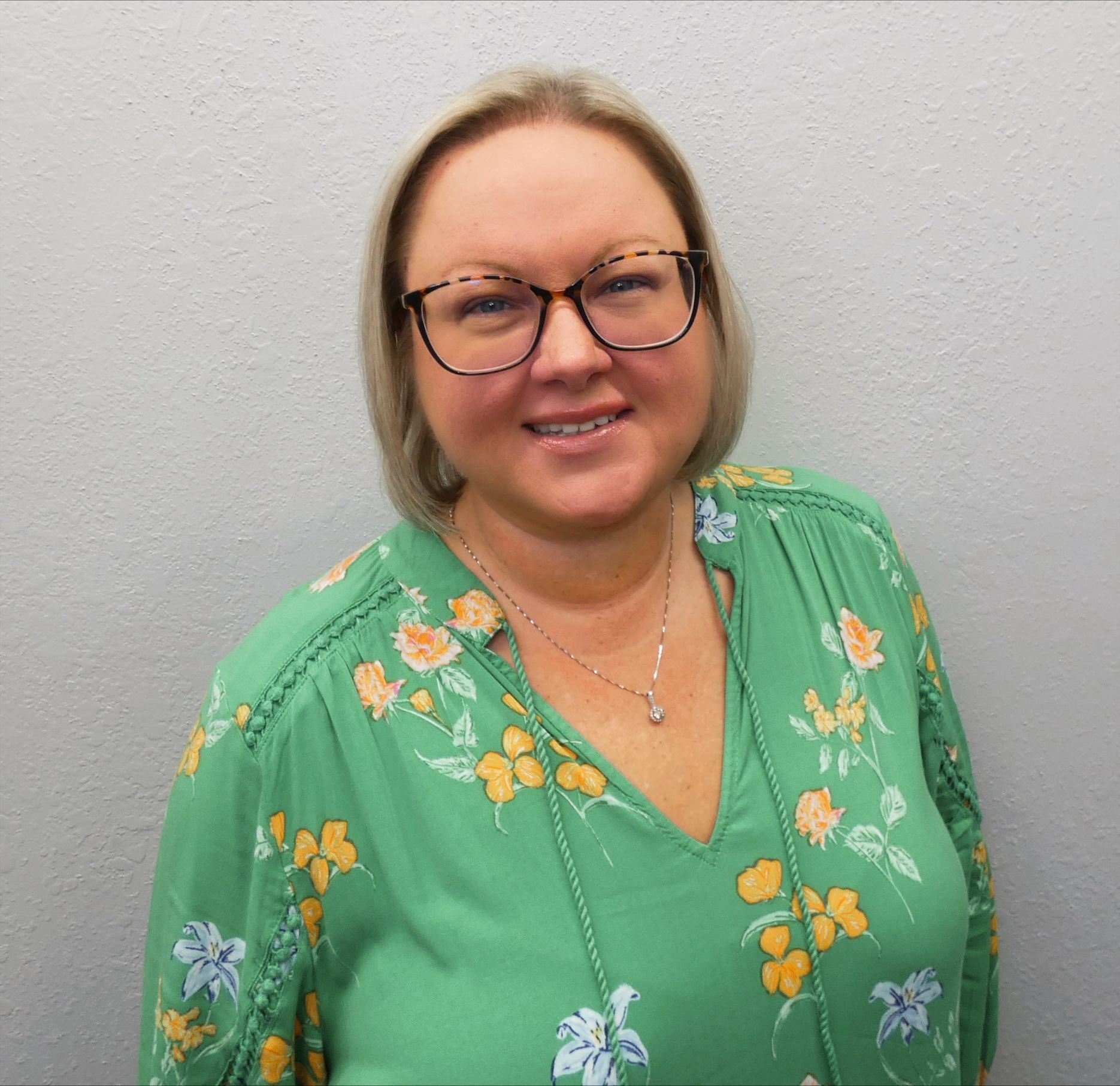 A woman wearing glasses and a green floral shirt is smiling for the camera.