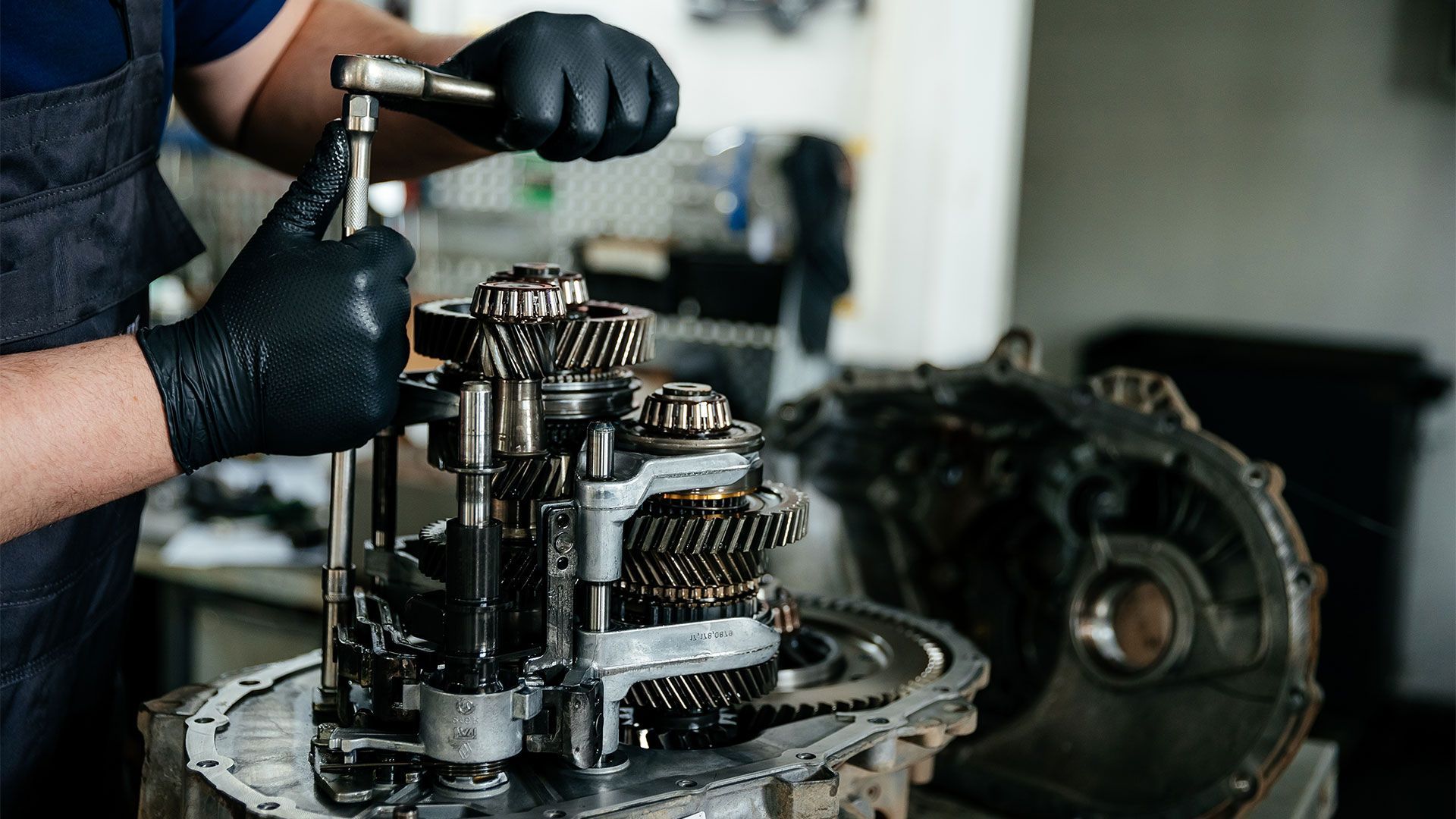 A man wearing black gloves is working on a gearbox in a garage.
