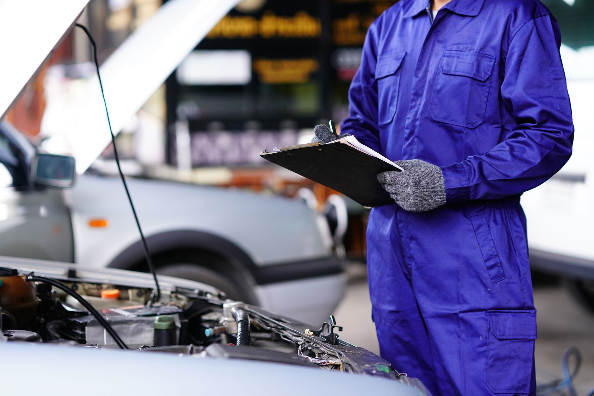 A mechanic is standing next to a car with the hood open and holding a clipboard.