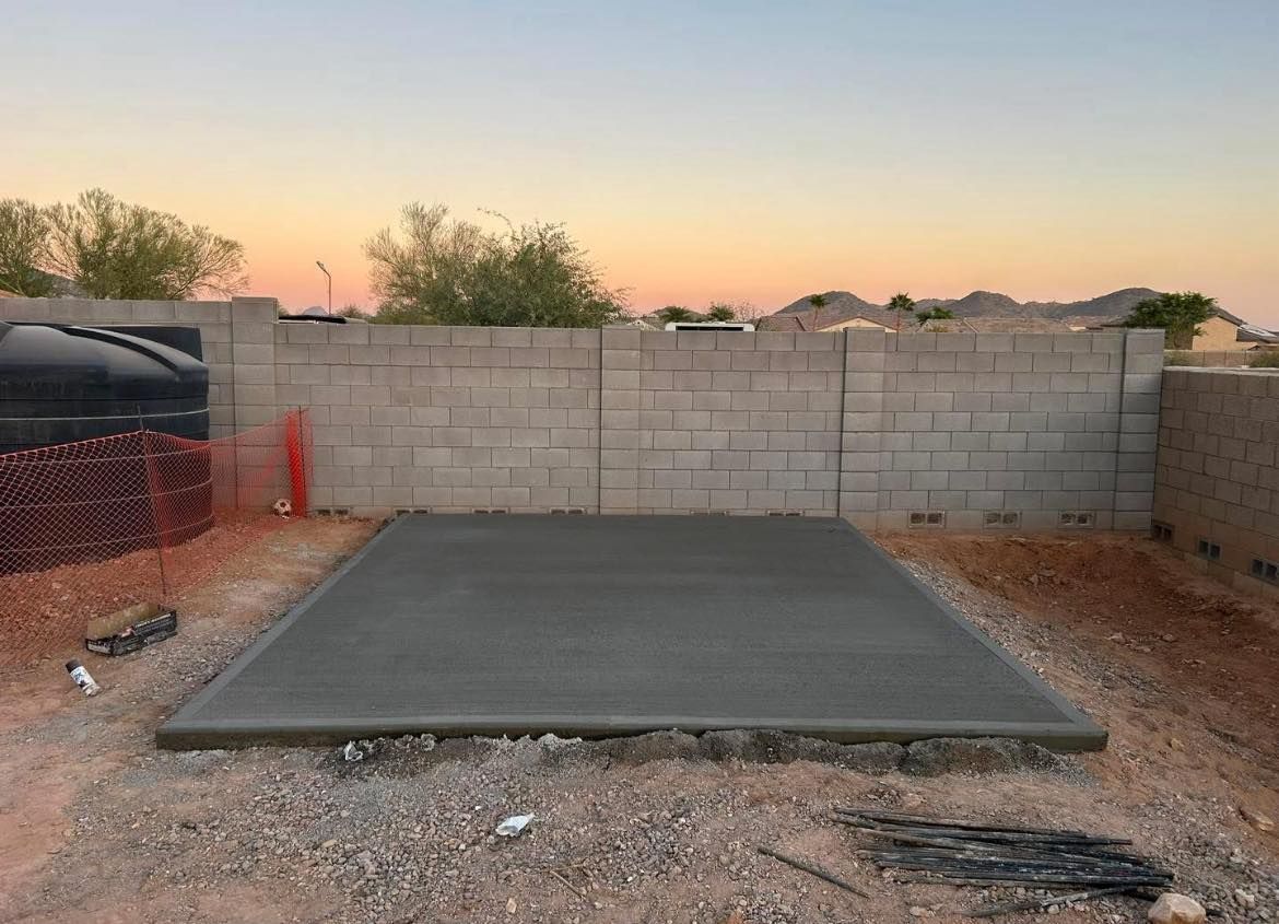 A freshly poured concrete pad in a backyard, surrounded by gravel and a block wall, under a dusky sky.