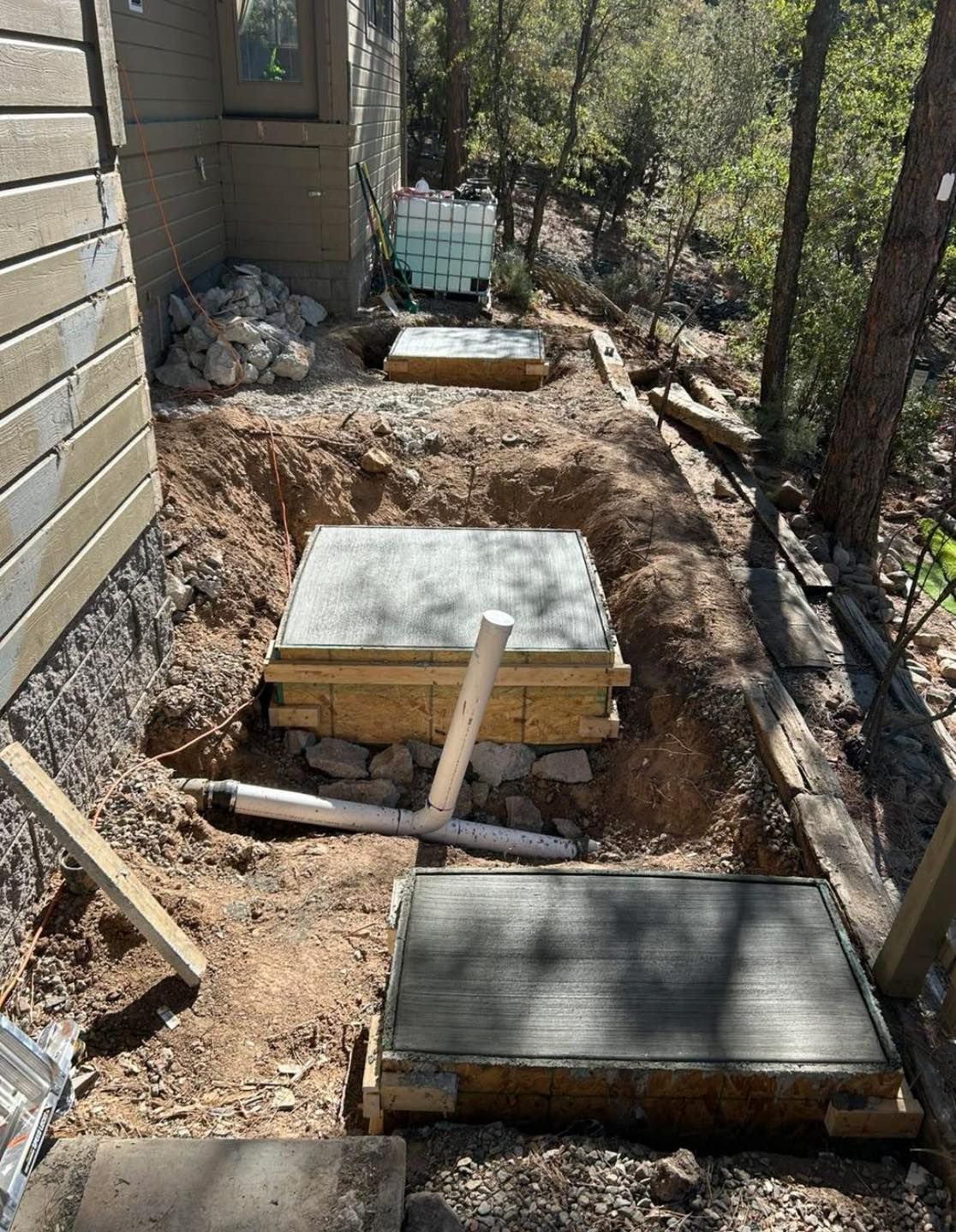 Concrete pads under construction along the side of a building on a dirt path.